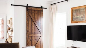 A living room with a brown sofa, a white armchair with a blue pillow, a wooden barn door, a television on a stand, and a painting on the wall. - Cozy Millbrook Cottage | Pool Access Near Capitol (Millbrook)
