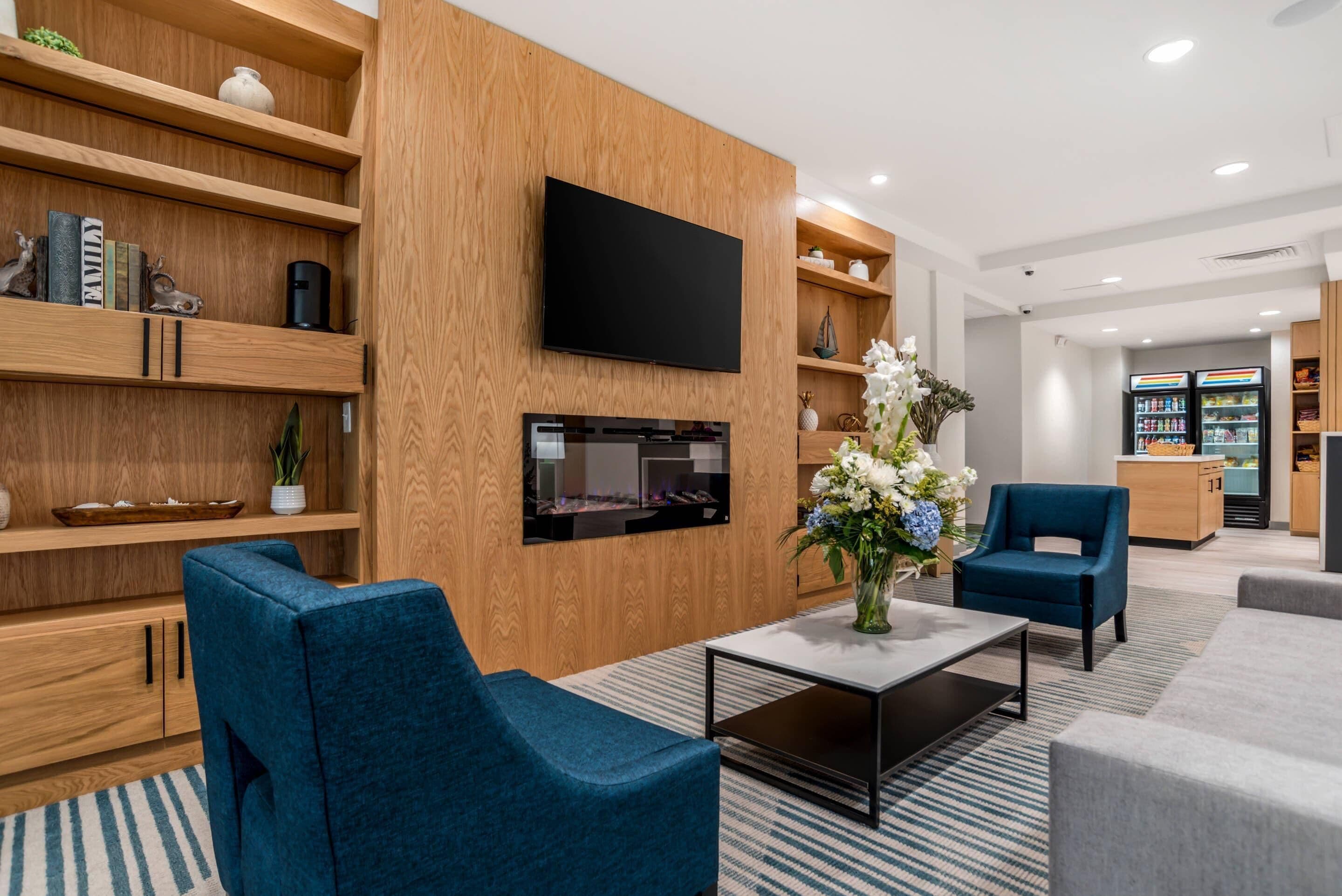 A modern hotel lobby with a flat-screen TV, wooden shelving with books and decorative items, a fireplace, and seating areas.