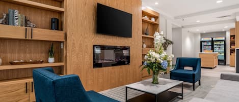 A modern hotel lobby with a flat-screen TV, wooden shelving with books and decorative items, a fireplace, and seating areas.