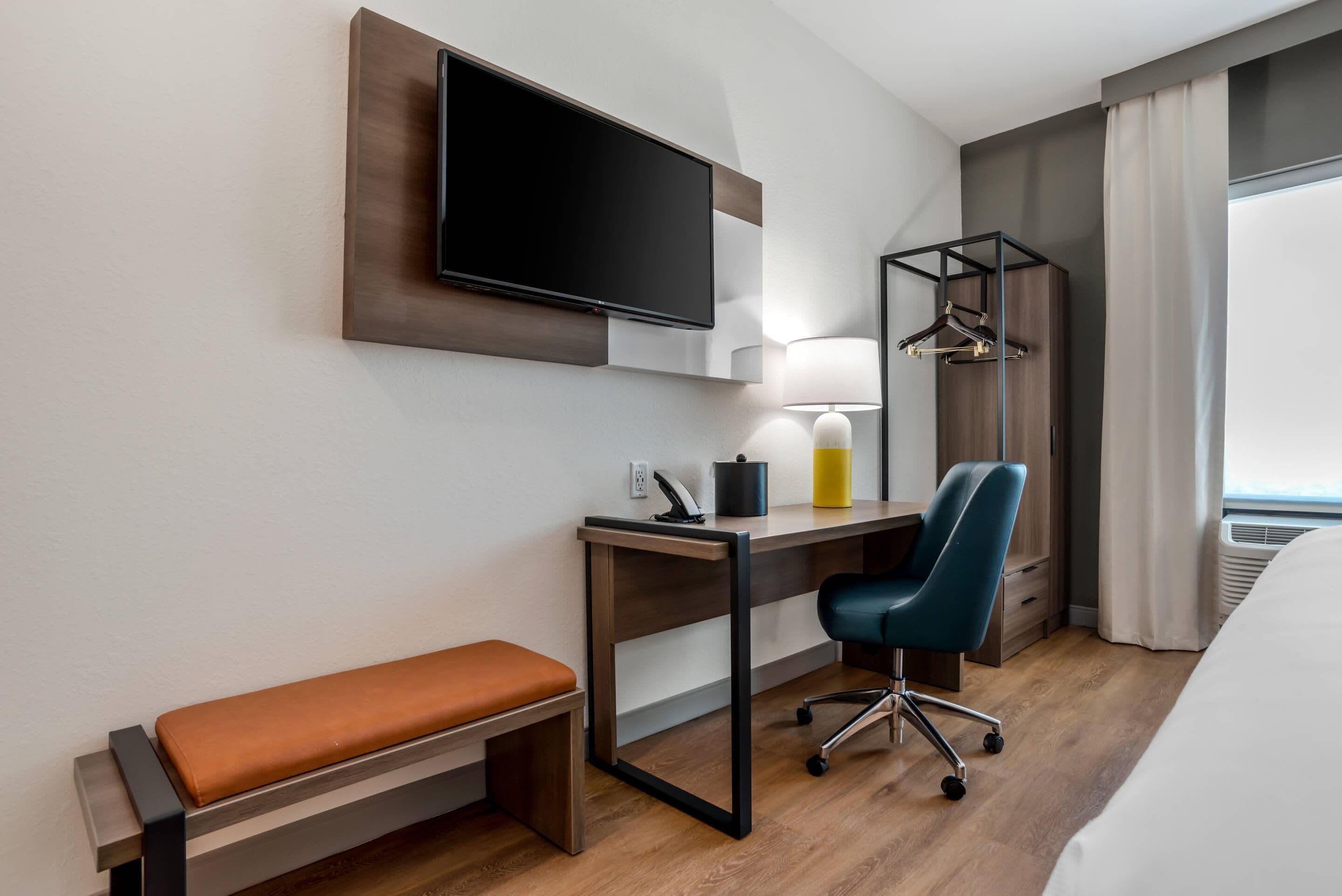 Una habitación de hotel con una cama, un escritorio con silla, un televisor, una lámpara y un perchero.
