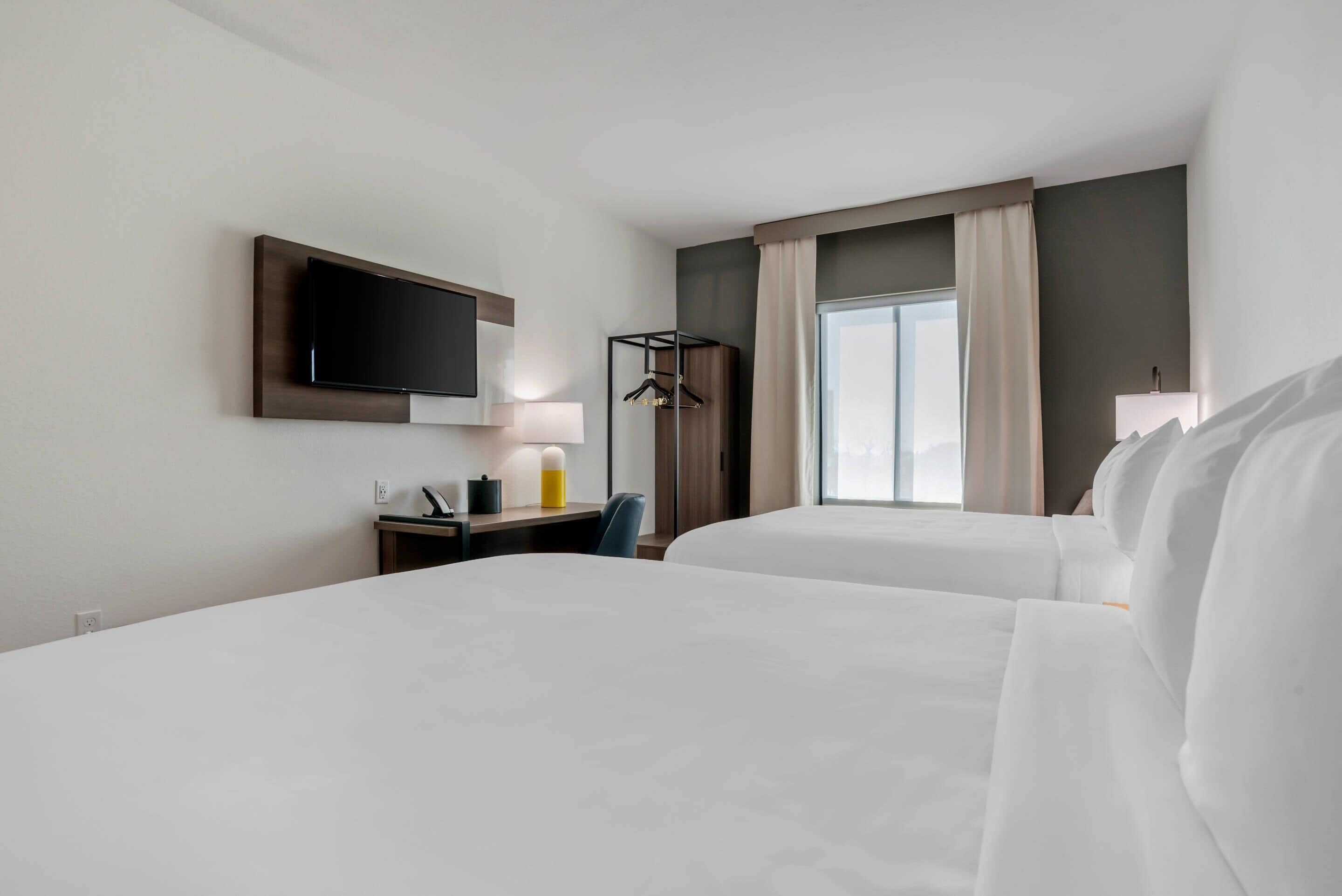 Een hotelkamer met twee bedden, een bureau met een lamp, een aan de muur bevestigde televisie en een raam met gordijnen.