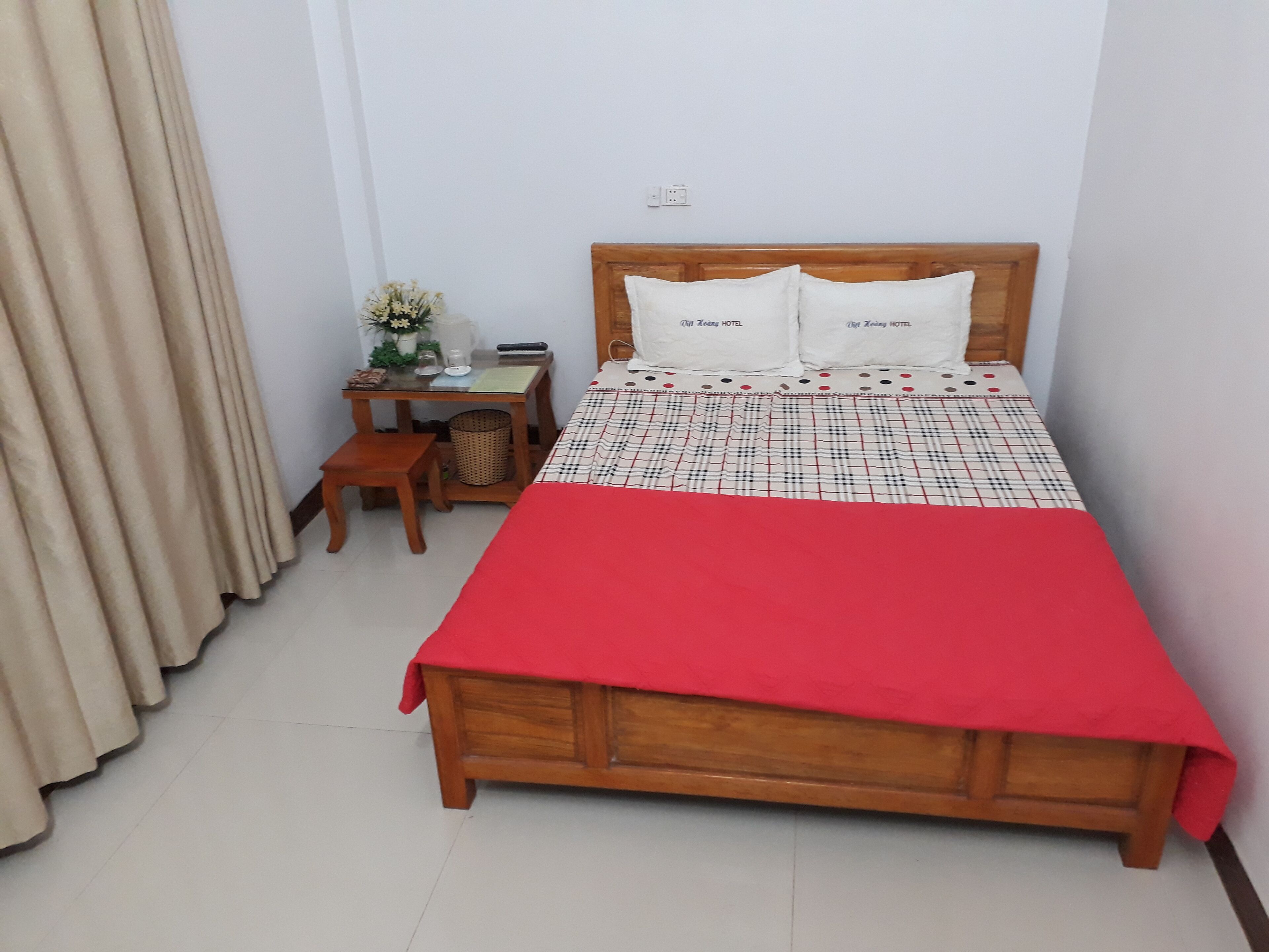 A neatly arranged hotel room with a wooden bed, a small wooden nightstand, a table with a vase of flowers, and a red bedspread.