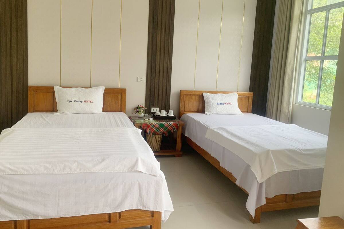 A hotel room with two single beds, a small table with a lamp, and a window with curtains.
