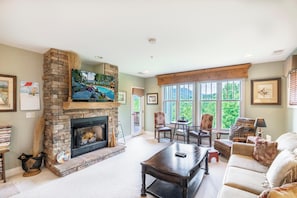 A living room with a stone fireplace, a flat-screen TV, a large window, and a cozy seating area. - Evergreens 1522- Mtn Condo, Gated Resort, Indoor/Outdoor Pools & Hot Tub (Boone)