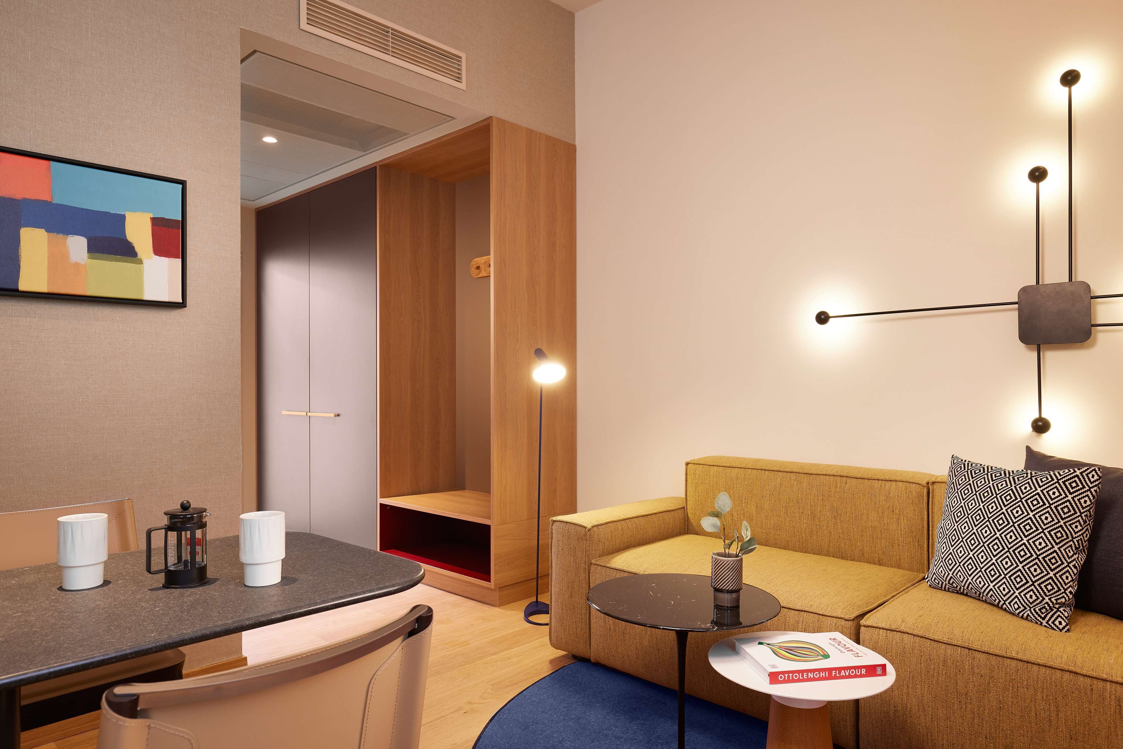 A modern hotel room with a wooden wardrobe, a sofa, a small table with a magazine, and a coffee maker on the counter.