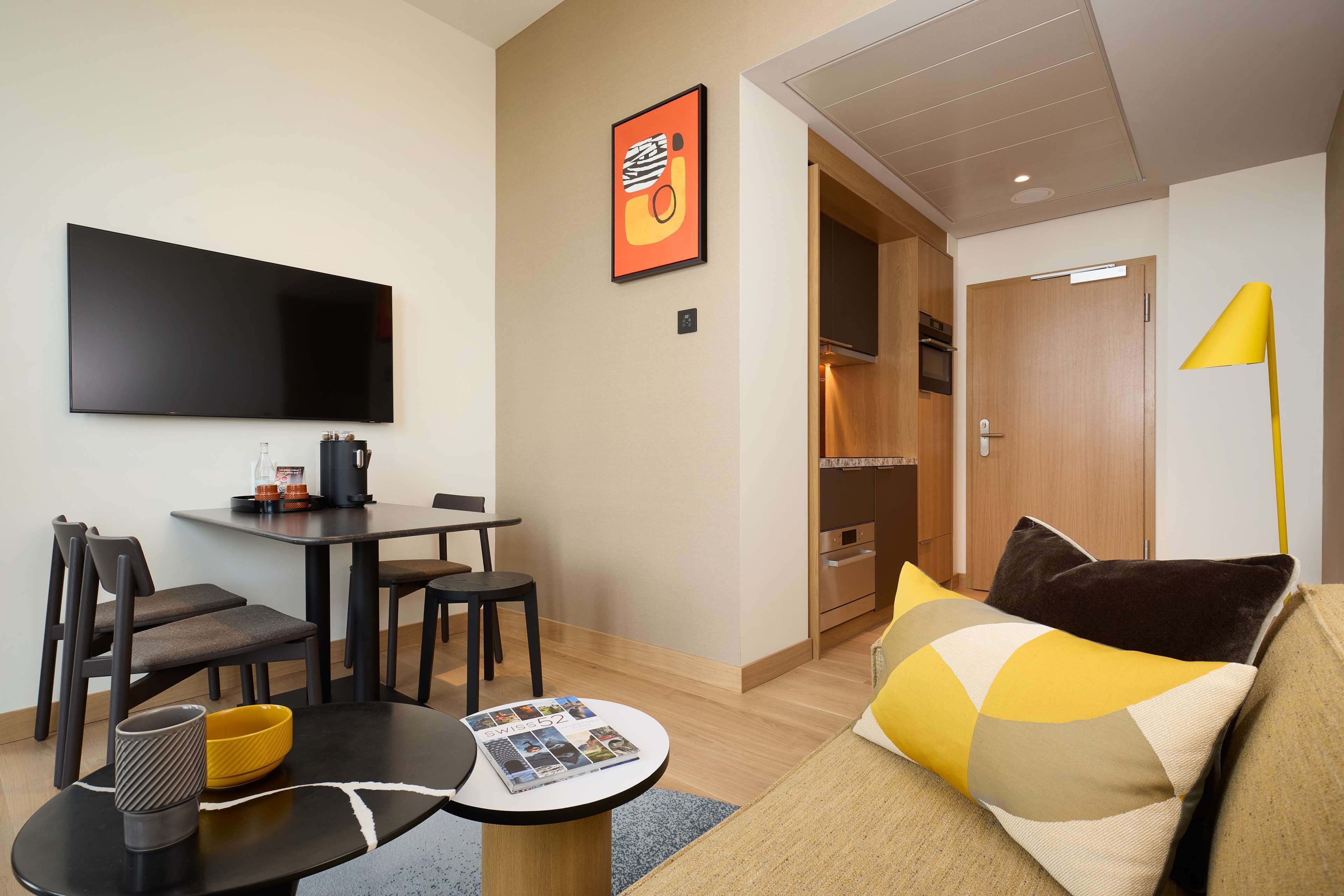 A modern living room with a flat-screen TV, a dining table with chairs, a sofa with cushions, and a yellow floor lamp.
