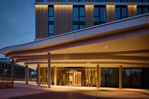 Una entrada de hotel moderna con un corredor cubierto, ventanas amplias y un llamativo letrero 'Adina'.
