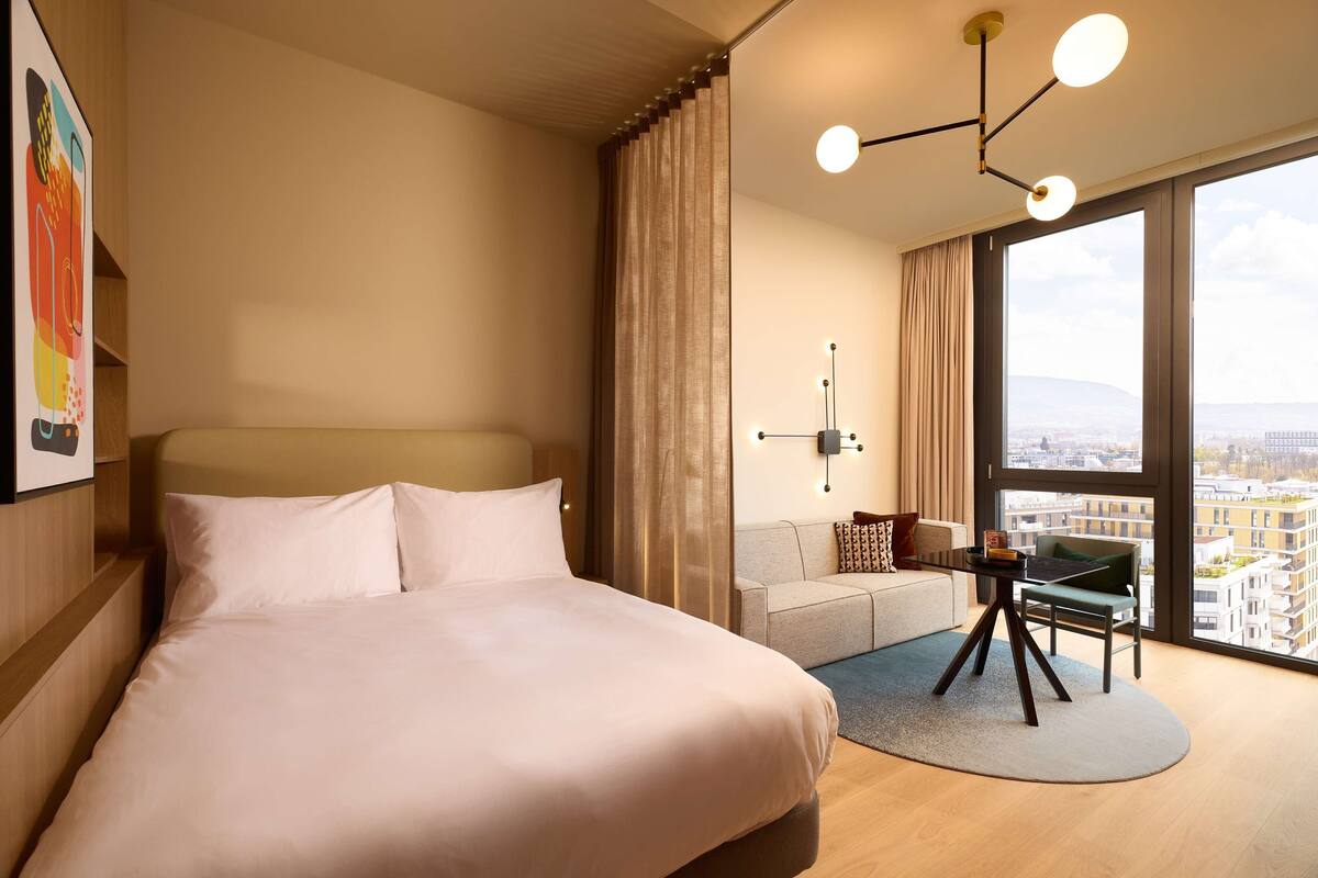 Habitación de hotel con una cama grande, un sofá, una mesa de comedor y vistas de la ciudad a través de amplios ventanales.