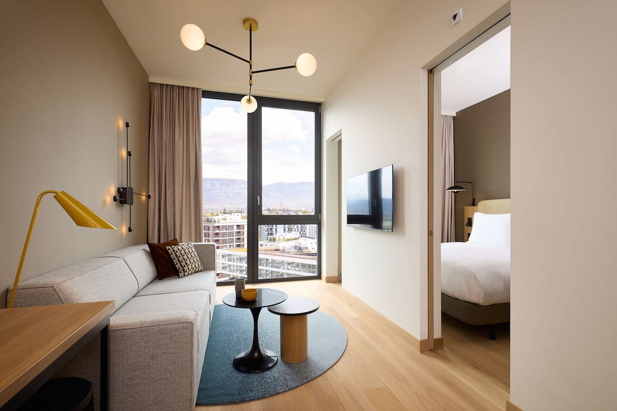 Habitación de hotel moderna con sofá, una mesita redonda, televisor y un amplio ventanal con vistas a la ciudad.
