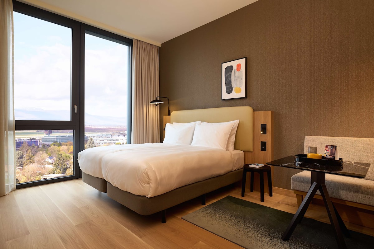 Una habitación de hotel moderna con una cama grande, un sofá, una mesita y vistas a la ciudad a través de un amplio ventanal.