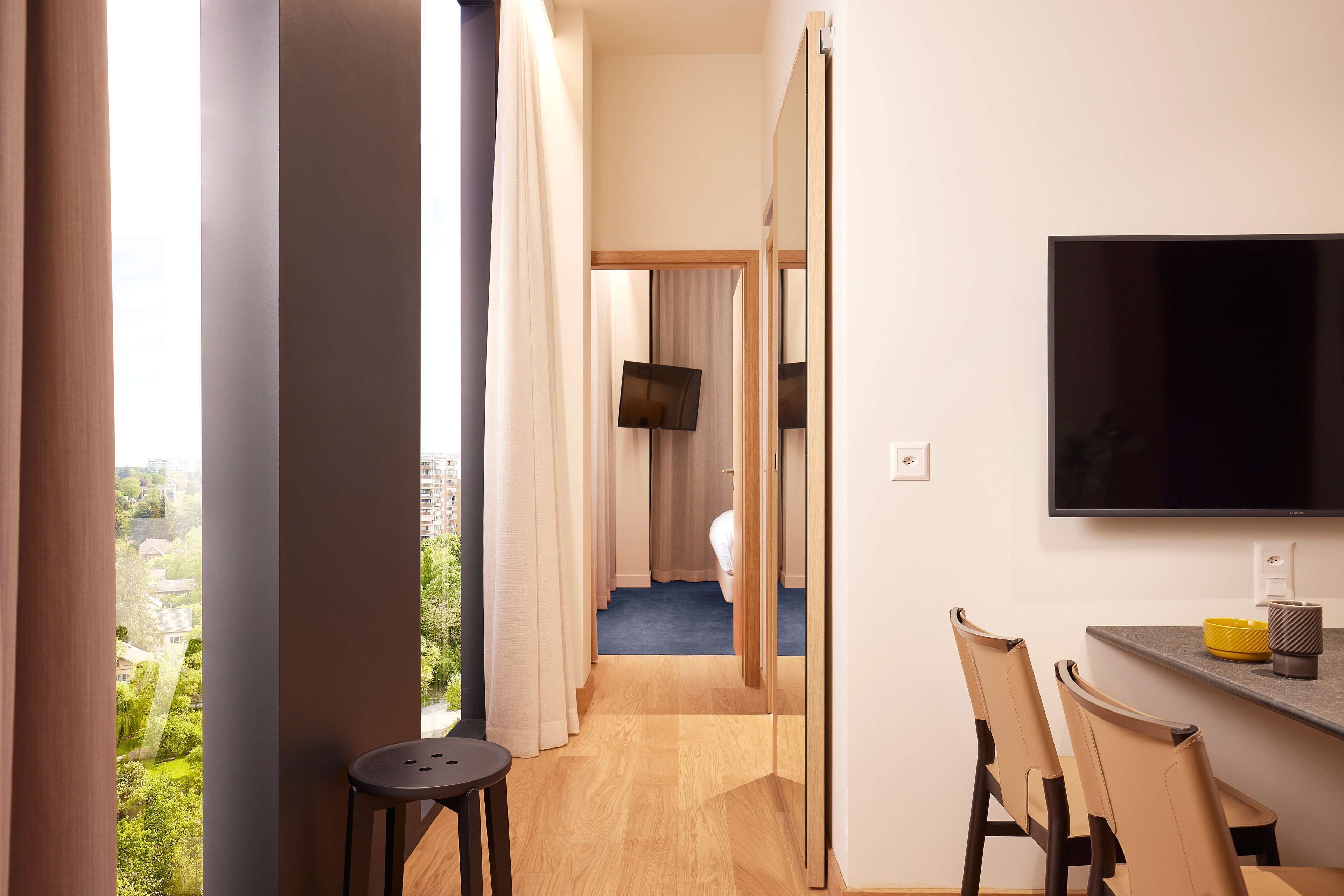 A modern hotel room with a large window, a flat-screen TV, a dining area with chairs, and a small stool.
