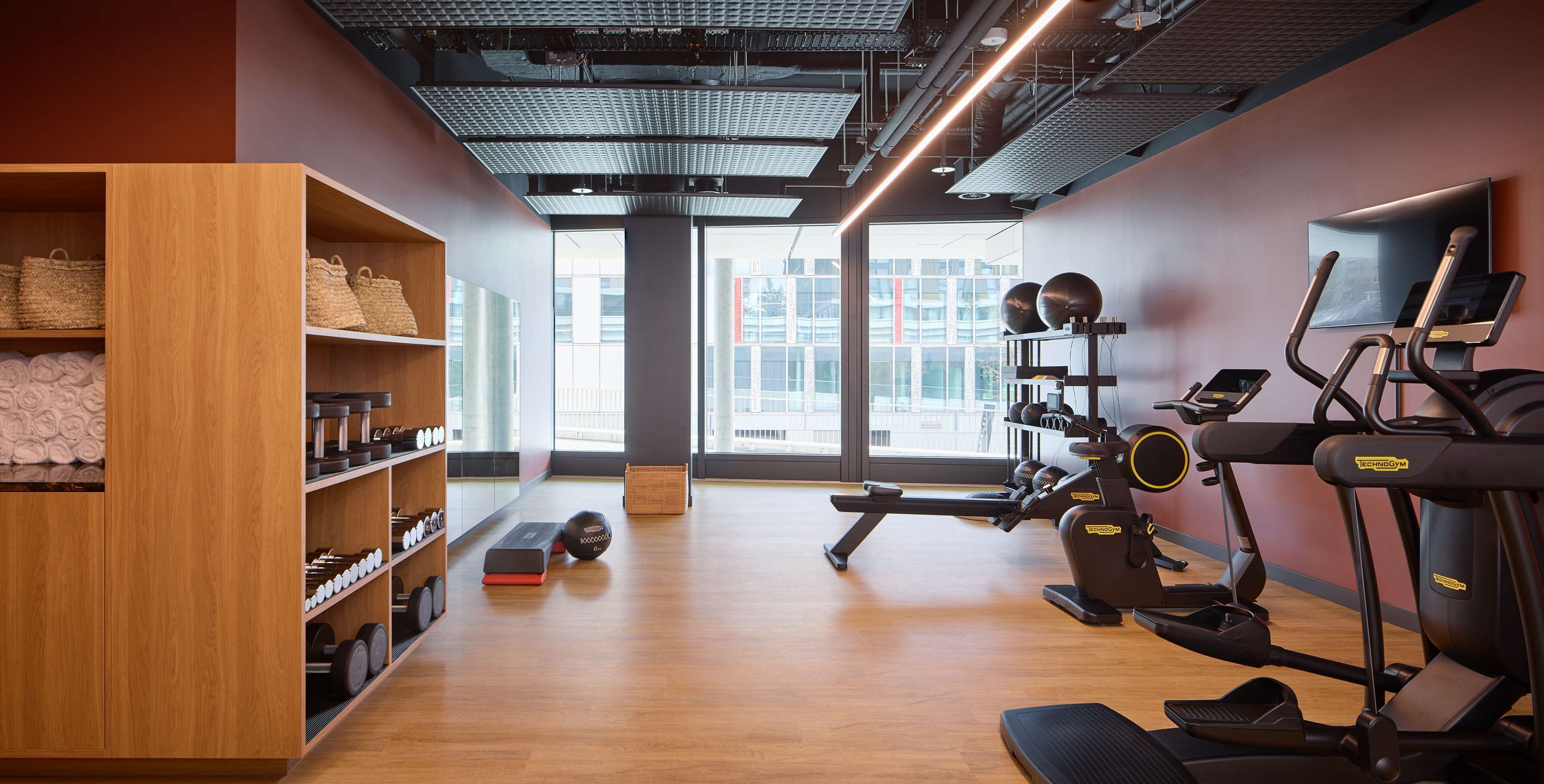 A modern gym with exercise equipment, wooden shelves with towels, and a large window offering a city view.