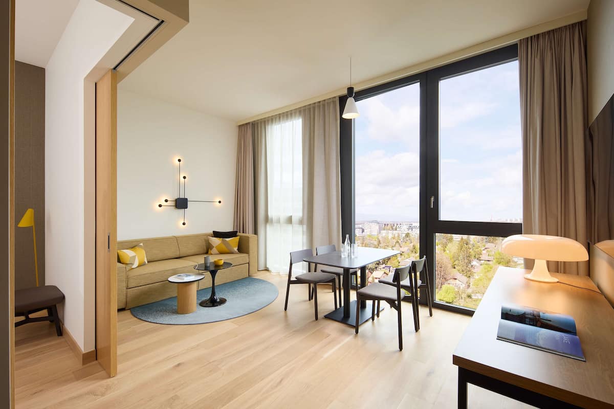 Una habitación de hotel moderna con un ventanal grande, una zona de comedor, un sofá y una lámpara.