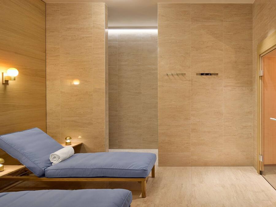 A modern spa room with wooden paneling, two blue cushioned chairs, a small table with a lamp, and a door with a sign.