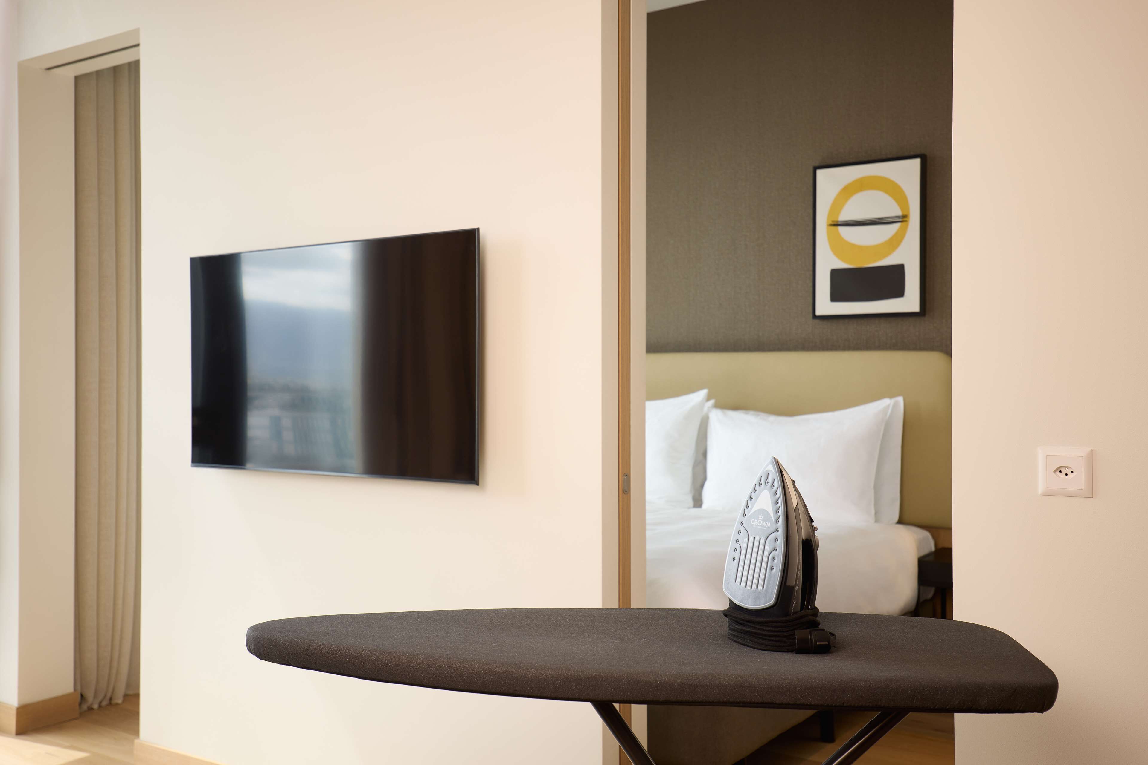 A hotel room with a flat-screen TV mounted on the wall, a bed with white bedding, and an ironing board with an iron.