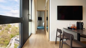 Een hotelkamer met een groot raam, een aan de muur bevestigde televisie, een bureau met stoelen en een bed met gordijnen.