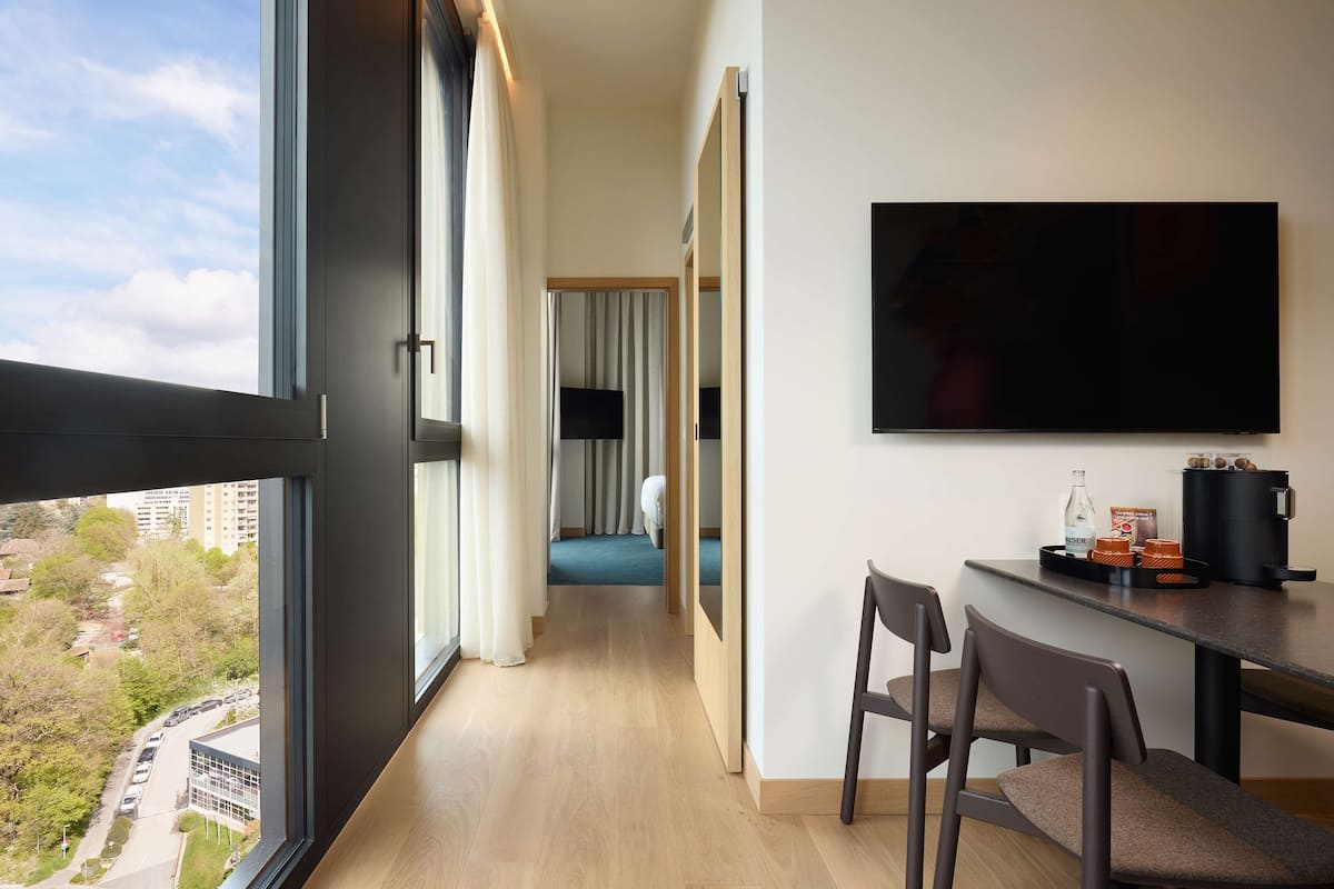 Habitación de hotel con un ventanal grande, televisor montado en la pared, escritorio con sillas y cama con cortinas.