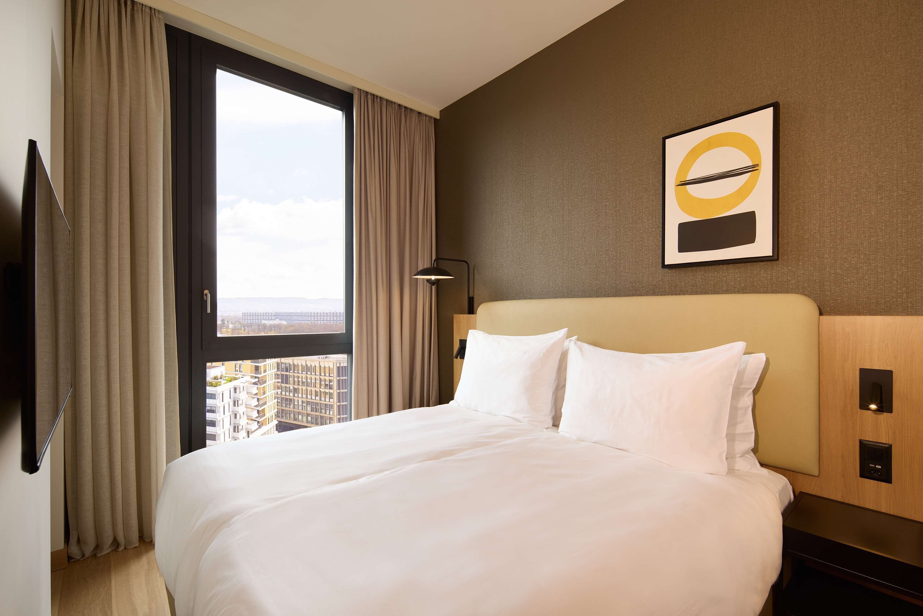 A hotel room with a large bed, a view of the city, a flat-screen TV, and a modern wall art piece.