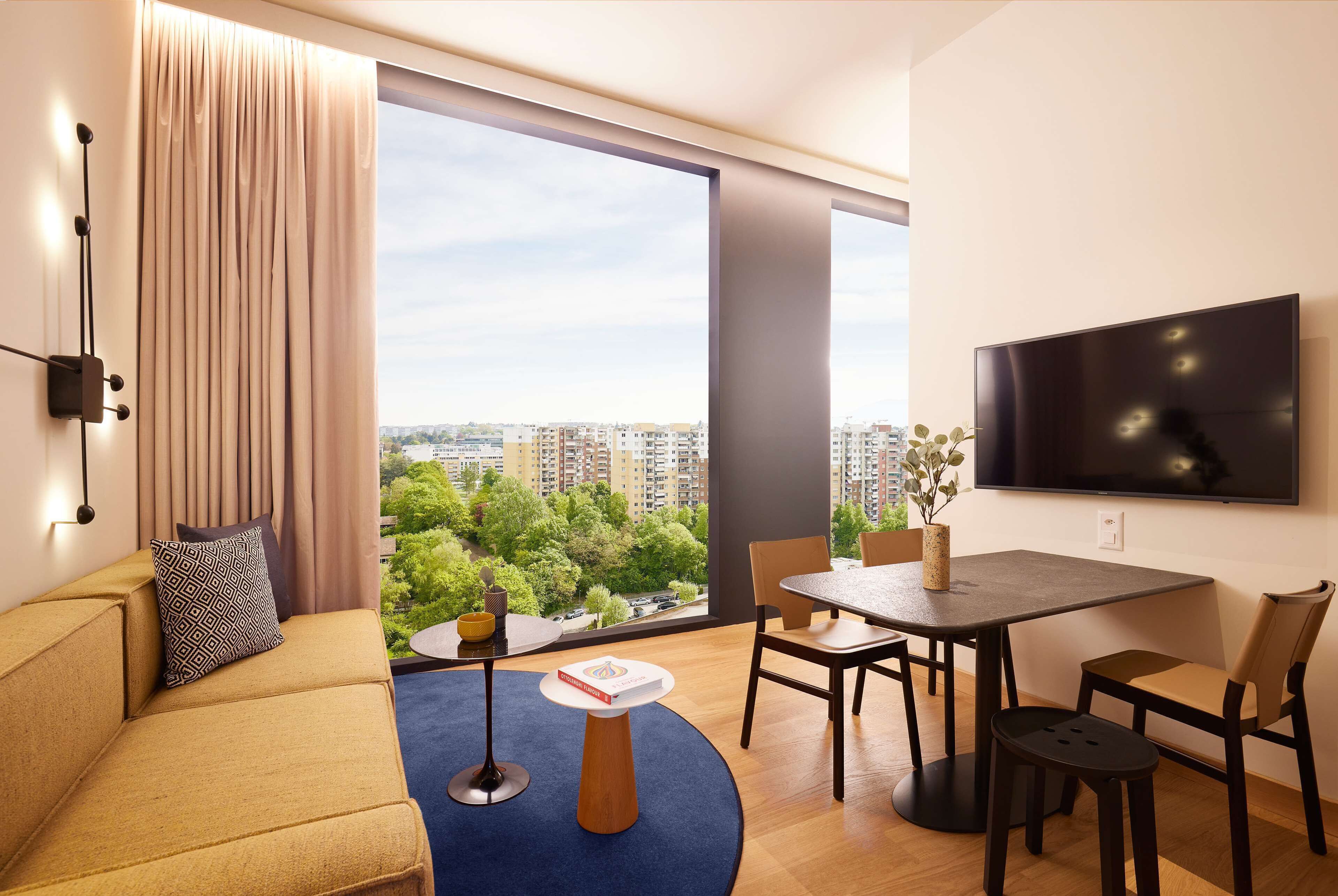 A modern living room with a sofa, a coffee table, a dining table with chairs, a flat-screen TV mounted on the wall, and a large window offering a city view.