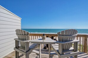 Um deck de madeira com duas cadeiras Adirondack e uma mesa, com vista para uma praia e oceano.