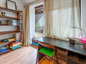 Een kamer met een houten bureau, een stoel met een groene kussentje, een boekenplank vol boeken en mappen, en een raam met gordijnen.