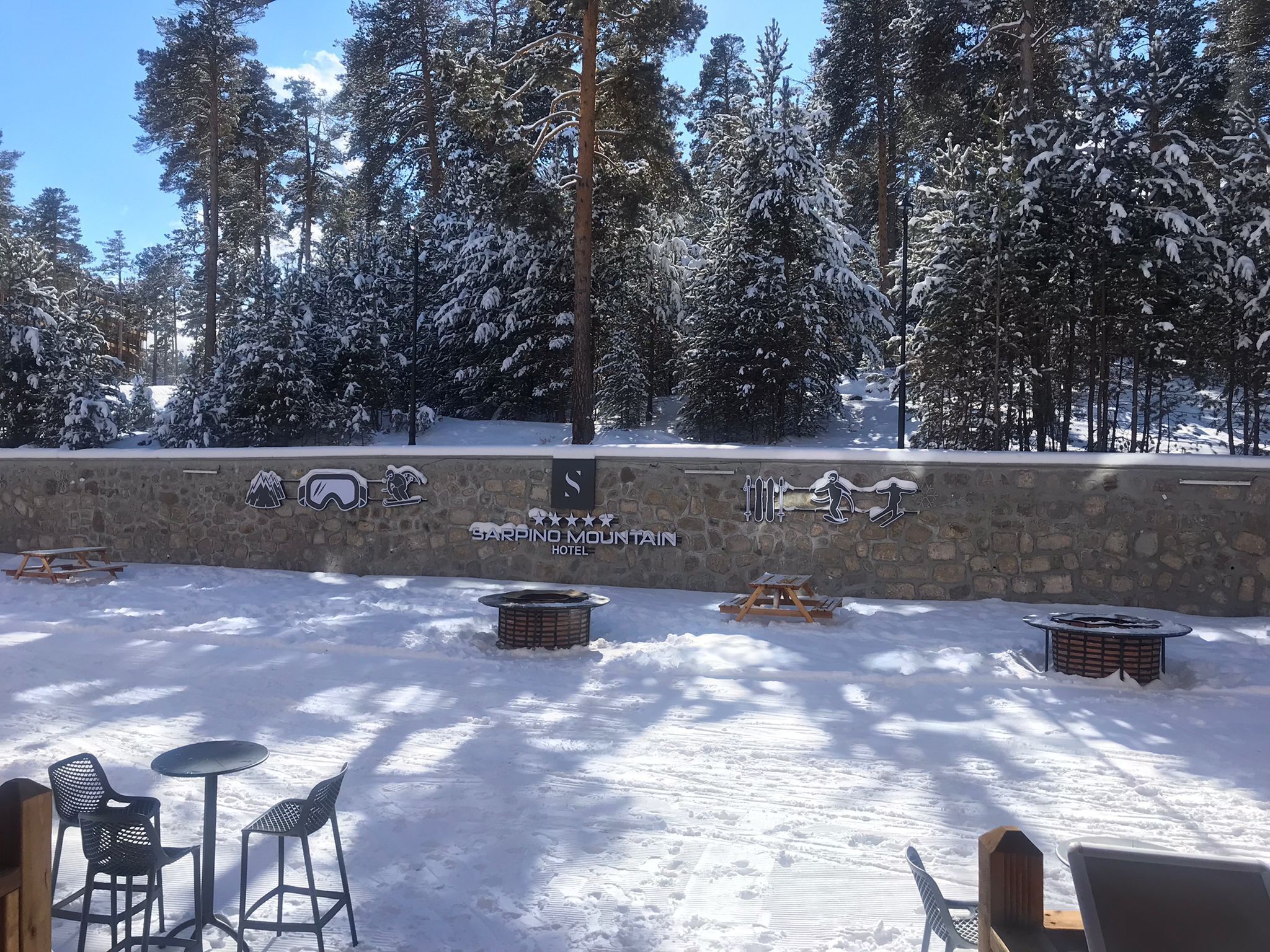 A snowy outdoor area with a stone wall displaying the Sarpino Mountain Hotel logo, picnic tables, and chairs.