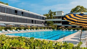 A swimming pool with lounge chairs and umbrellas, surrounded by greenery and modern buildings. - Bilo Standard by Interhome (Marina di Bibbona)