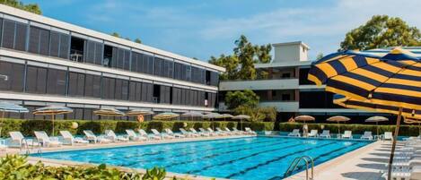 A swimming pool with lounge chairs and umbrellas, surrounded by greenery and modern buildings.