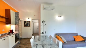 Une cuisine moderne avec une cuisinière à gaz, des armoires blanches et un mur d’accent en pierre. On y trouve une table à manger avec des chaises, ainsi qu’un canapé gris agrémenté de coussins orange.