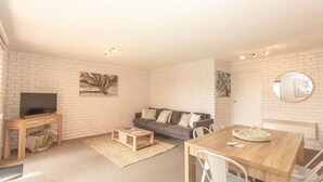 A modern living room with a flat-screen TV, a grey sofa, a wooden coffee table, and a dining table with chairs. - Sunrise - 15/121 Gippsland Street (Jindabyne)
