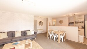 A modern living room with a grey sofa, a wooden coffee table, and a dining area with a wooden table and white chairs. - Sunrise - 15/121 Gippsland Street (Jindabyne)