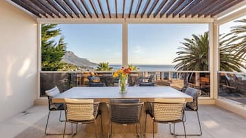 A terrace with a table and chairs, overlooking a coastal landscape with vegetation and a mountain.