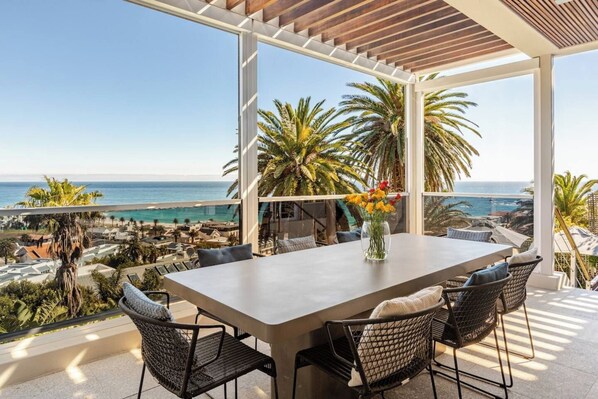 A terrace with a table and chairs, overlooking a coastal view with palm trees and a clear sky.