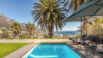 Une piscine avec une terrasse en bois, des chaises longues et une vue sur l’océan et les montagnes.