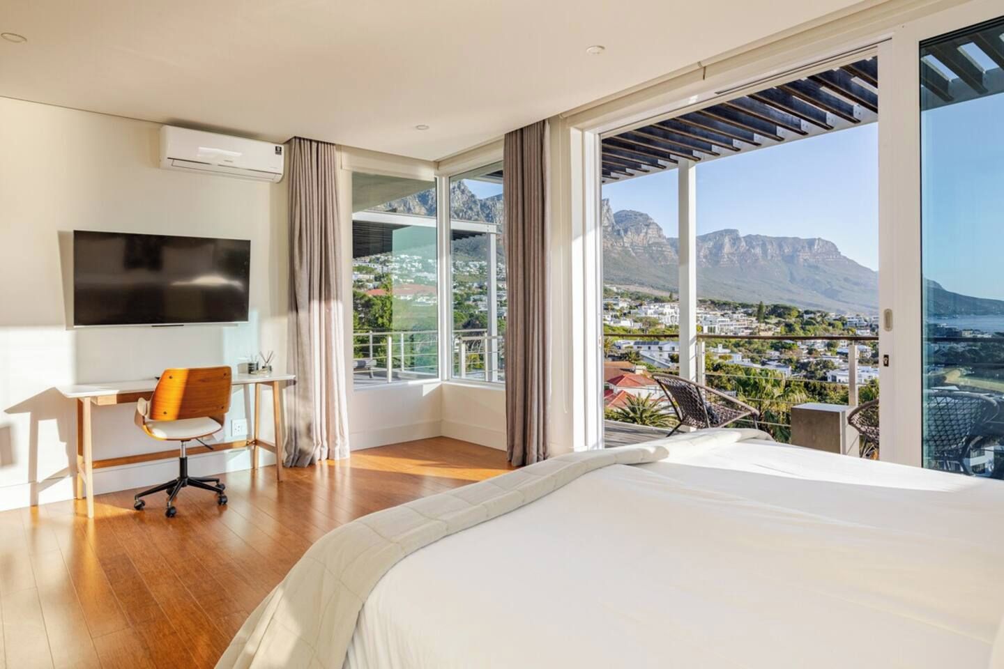 A modern bedroom with a large bed, a desk, and a TV. It has a balcony with a city view.