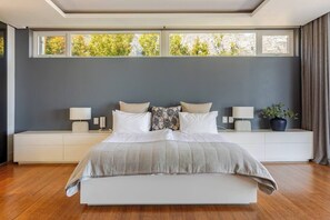 A modern bedroom with a large bed, bedside tables, and a view of trees through the windows.