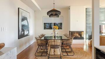 A modern dining area with a glass table, wicker chairs, a central vase with flowers, and a fireplace in the background.