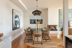 A modern dining area with a glass table, wicker chairs, a central vase with flowers, and a fireplace in the background.