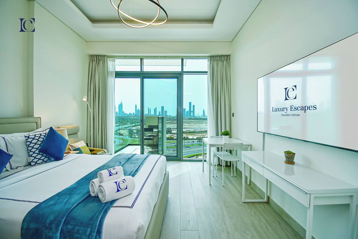 A hotel room with a large bed, a desk, a chair, and a TV displaying the hotel name.