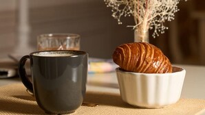 Une table avec une tasse noire, un verre de café, un bol blanc contenant un croissant et un vase avec des fleurs séchées.