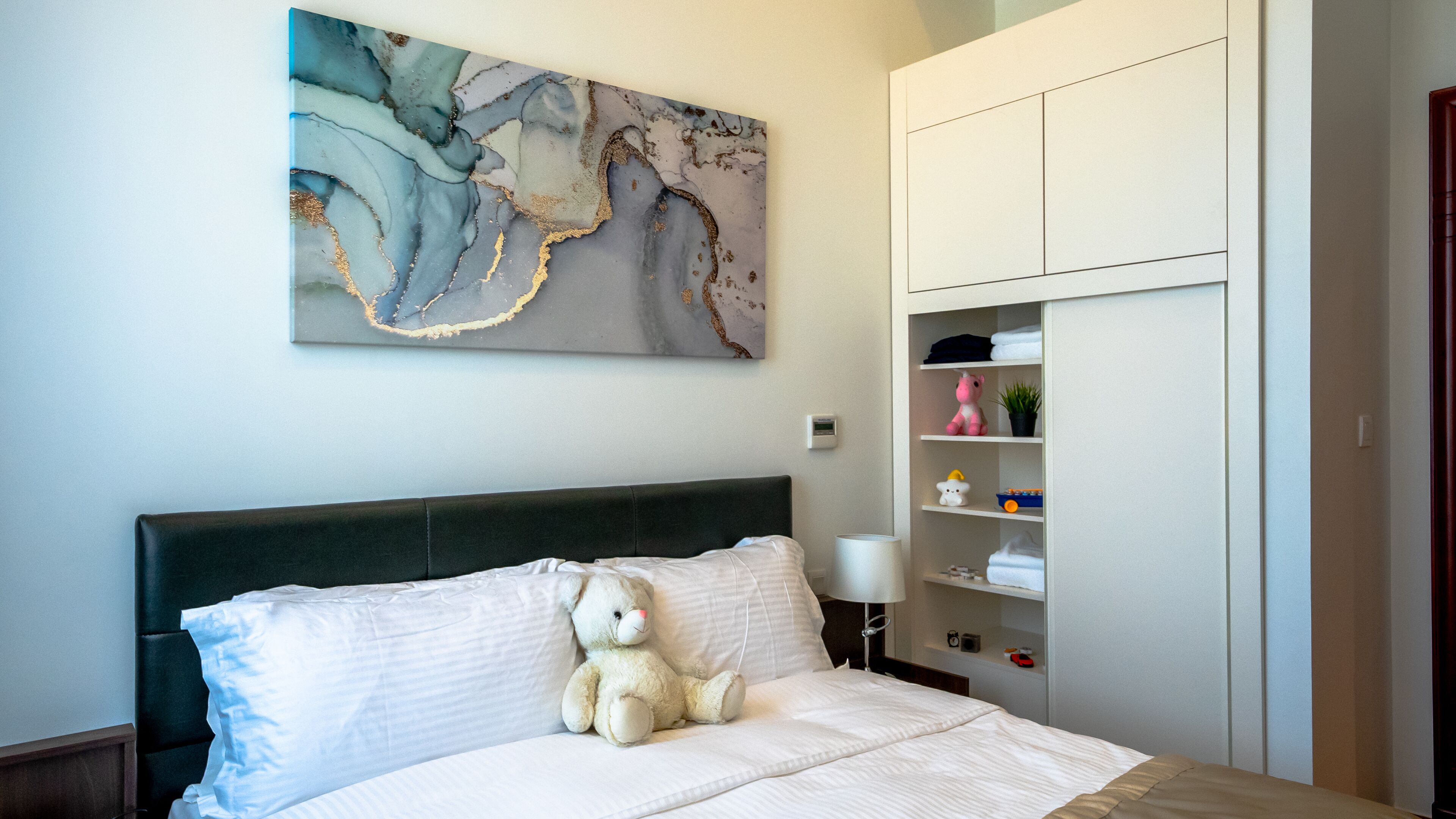 A bedroom with a bed, a teddy bear, a nightstand, a lamp, a wall-mounted artwork, and a built-in wardrobe.