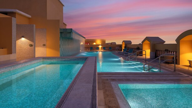 Zona de piscina con agua cristalina, solárium con baldosas y cielo al atardecer.