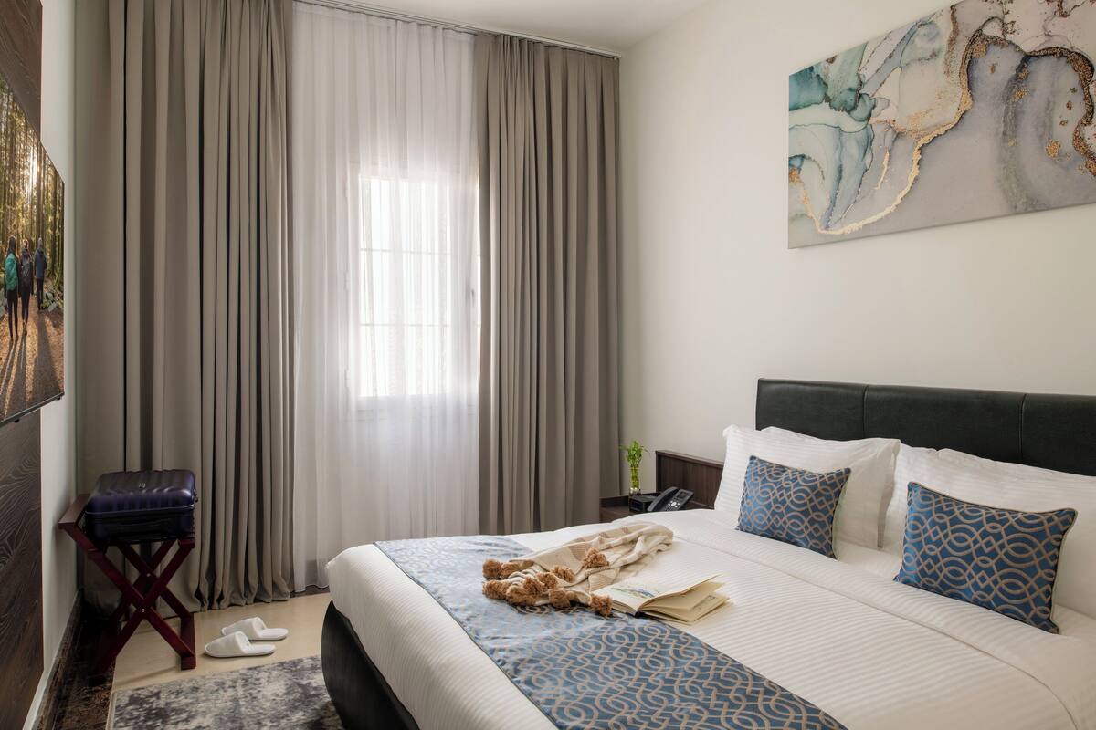 A hotel room with a bed, curtains, a small table with a suitcase, slippers, a bedside table with a phone, and a wall art piece.