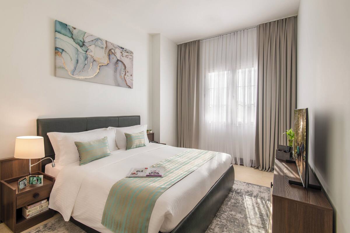 A neatly arranged hotel room with a large bed, a bedside table with a lamp, a flat-screen TV, and a painting on the wall.