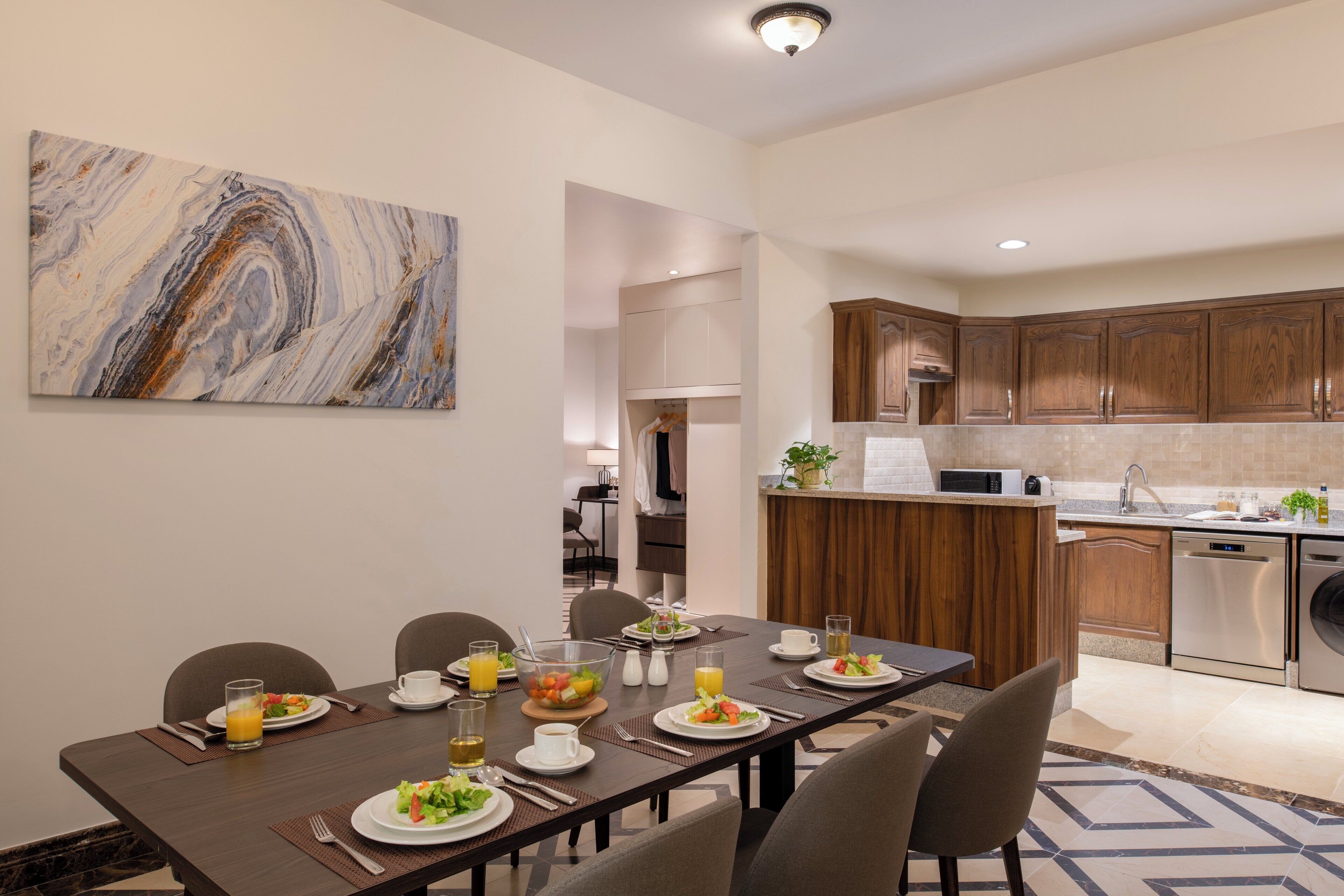A modern dining area with a long table set for a meal, a kitchen with wooden cabinets, and a marble artwork on the wall.
