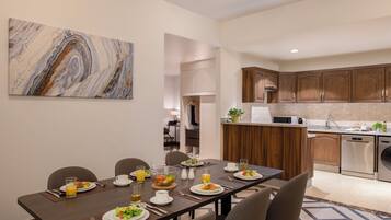 A modern dining area with a long table set for a meal, a kitchen with wooden cabinets, and a marble artwork on the wall.