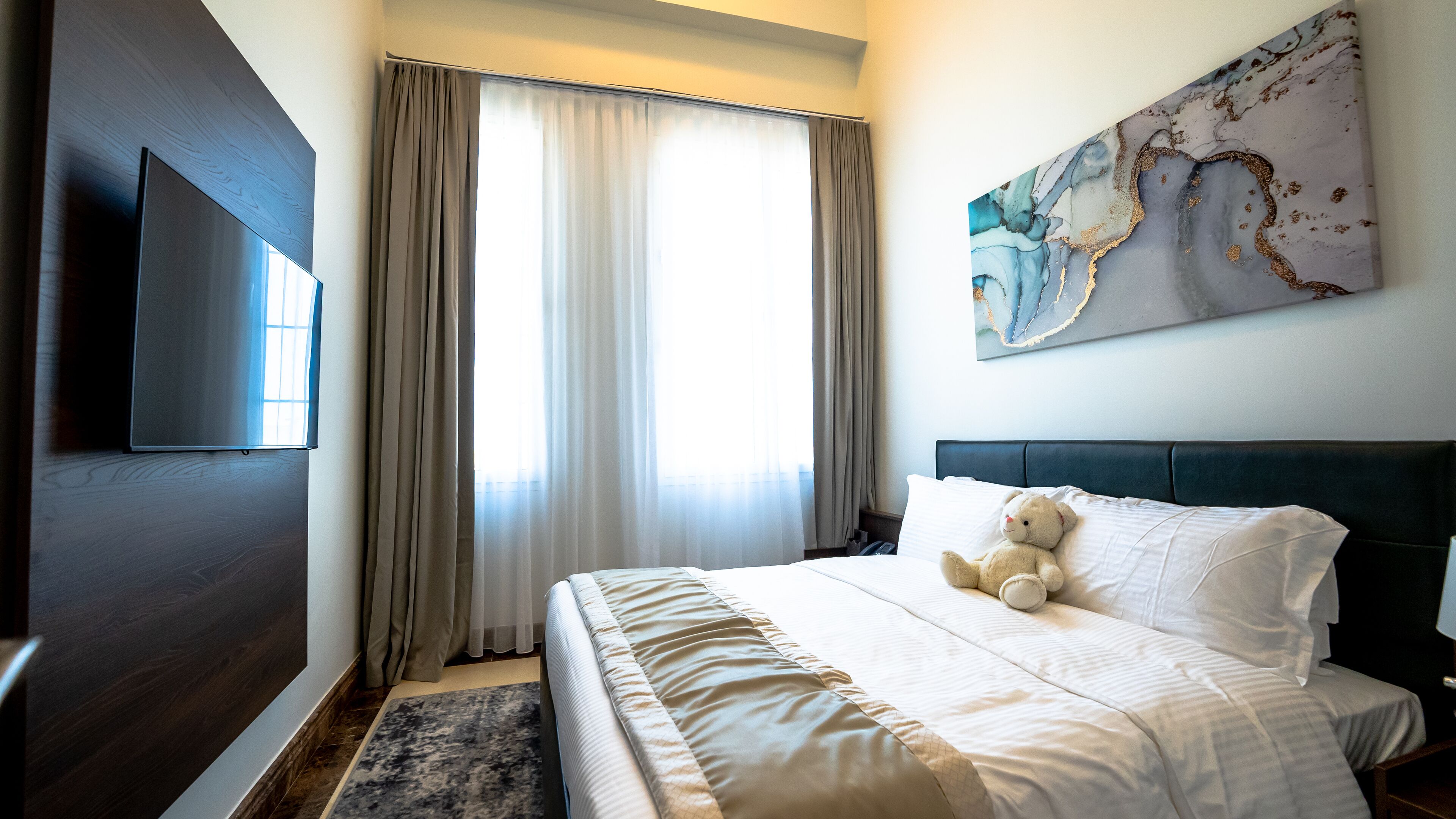 A hotel room with a bed, a television, a window with curtains, and a painting on the wall.