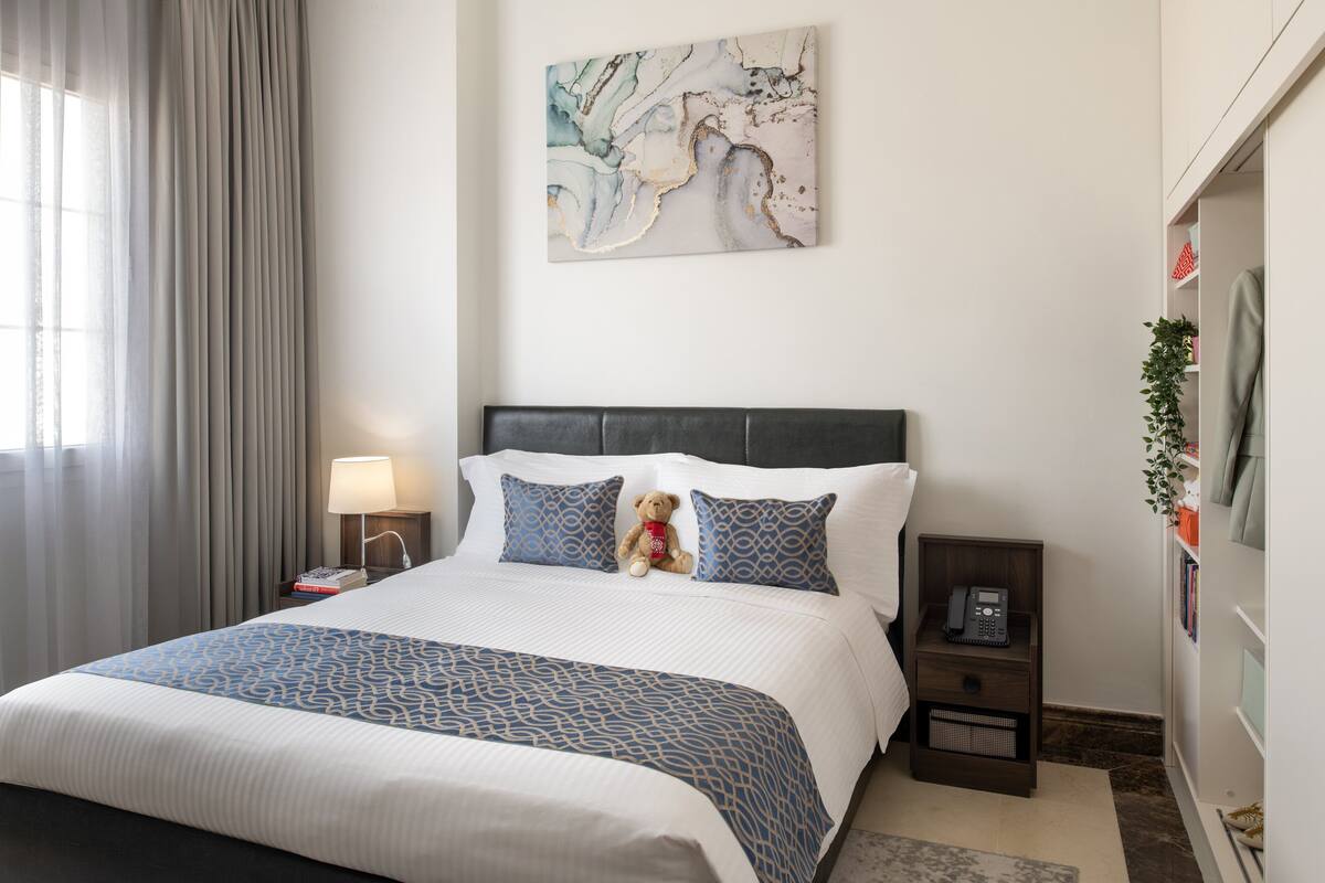 A neatly made bed with a patterned bedspread, a bedside lamp, a teddy bear, and a wall-mounted artwork.