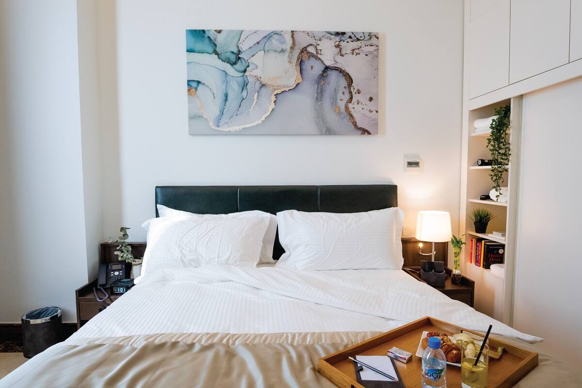 A hotel room with a bed, a bedside table with a lamp, a tray with a bottle and glasses, and a wall-mounted abstract painting.