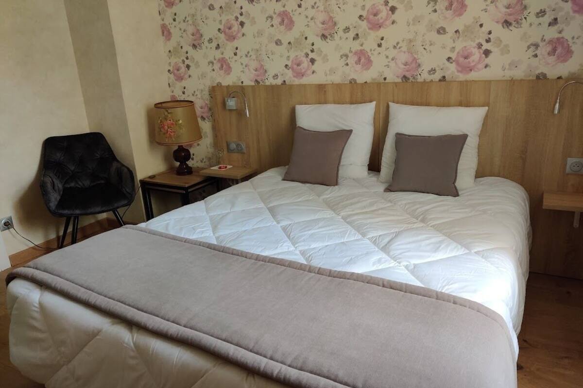 Uma cama bem arrumada com colcha bege e almofadas, uma cabeceira de madeira, uma pequena mesa de cabeceira com um abajur e papel de parede floral.